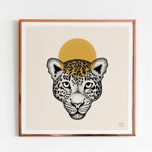 May include: A framed art print featuring a detailed illustration of a jaguar's face. The jaguar's fur is depicted with black spots and a yellow-gold sun shape is behind the head. The artwork is set against a cream background.