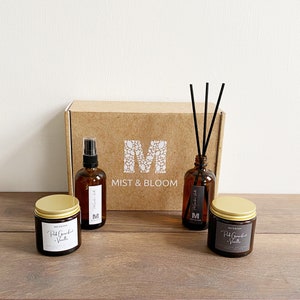 May include: A brown cardboard box with the text "Mist & Bloom" and a white letter "M" logo. The box contains three brown glass bottles with labels. Two bottles are candles and one bottle is a diffuser. The diffuser has black reeds.