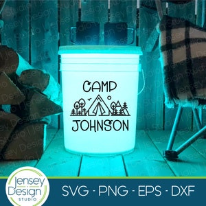 May include: A white plastic bucket with a blue glow, featuring a black silhouette of a tent, trees, and mountains with the text "CAMP JOHNSON" on it.