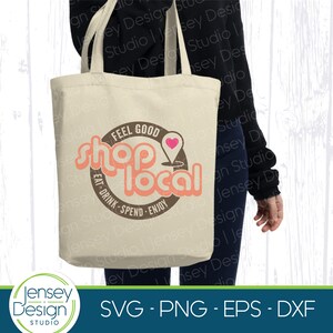 May include: A white tote bag with a brown circle design that says "Feel Good Shop Local Eat Drink Spend Enjoy" with a pink heart icon. The bag is being held by a person wearing a black shirt.
