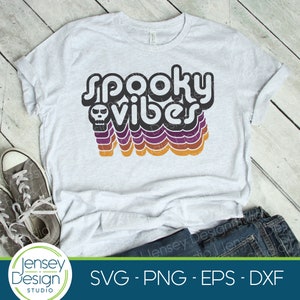 Puede incluir: Una camiseta blanca con un gr&aacute;fico negro que dice "spooky vibes" en una fuente retro. El gr&aacute;fico incluye una calavera y rayas de arco&iacute;ris en tonos naranja, morado y negro.
