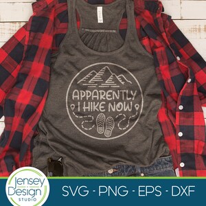 May include: A gray tank top with a white circle design featuring mountains, the text "APPARENTLY I HIKE NOW", and footprints. The tank top is laid out on a white wooden surface with a red and black plaid shirt and blue jeans.