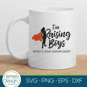 May include: White ceramic mug with a black silhouette of a woman wearing a red cape and the text "I'm Raising Boys" in black. The text "What's your superpower?" is also printed in black below the main text.