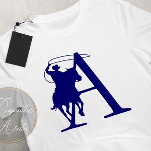 May include: A white t-shirt with a blue silhouette of a cowboy riding a horse and lassoing a rope. The silhouette is in the shape of the letter "A".