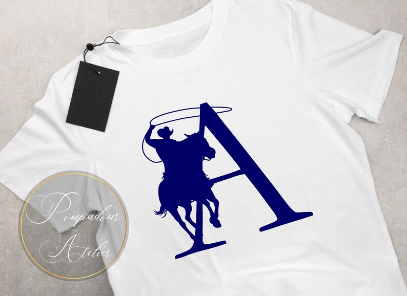 May include: A white t-shirt with a blue silhouette of a cowboy riding a horse and lassoing a rope. The silhouette is in the shape of the letter "A".