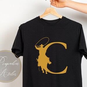 May include: Black t-shirt with a gold silhouette of a cowboy riding a horse and lassoing a rope, forming the letter "C".