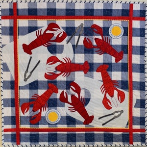 May include: A white and blue checkered fabric with red accents features an embroidered design of five red lobsters and two silver lobster crackers. The lobsters are arranged in a circular pattern with their tails pointing outwards.