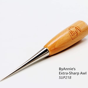 May include: A light brown wooden awl with a polished metal tip. The handle has a logo and text that reads "ByAnnie's Extra-Sharp Awl SUP218". This tool is designed for piercing and marking materials.