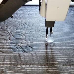 May include: A close-up of a longarm quilting machine in operation, stitching a pattern of swirling lines on a dark gray fabric.