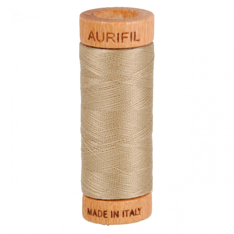 May include: A spool of Aurifil thread in a light beige color, wound on a wooden bobbin with the brand name "Aurifil" printed on the top and "Made in Italy" printed on the bottom.