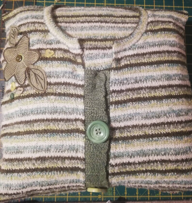 Puede incluir: Una funda de almohada a rayas hecha de un su&eacute;ter reutilizado con un bot&oacute;n verde y un adorno floral.