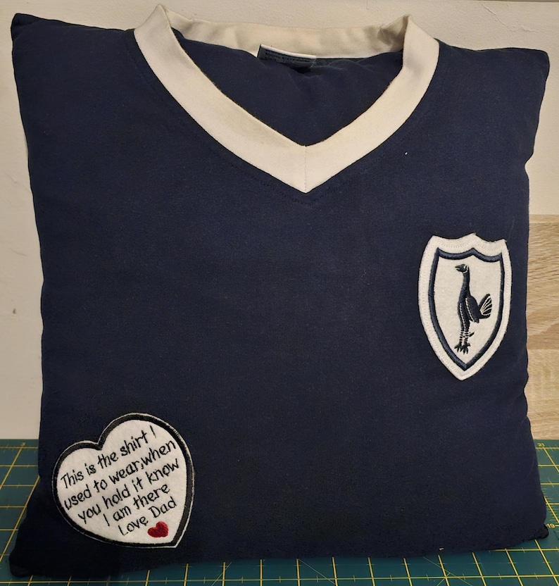 Puede incluir: Una almohada azul marino con cuello en V blanco y un escudo bordado blanco con un p&aacute;jaro. La almohada tiene un parche en forma de coraz&oacute;n con el texto "This is the shirt I used to wear, when you hold it know I am there Love Dad".