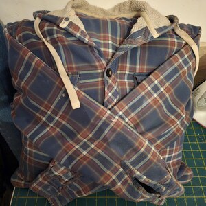 Puede incluir: Una camisa de franela a cuadros azules y rojos con capucha forrada de polar. Las mangas est&aacute;n remangadas y la camisa est&aacute; doblada por la mitad.