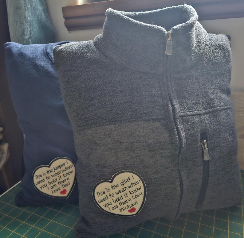 Puede incluir: Dos prendas de punto de polar gris con parches en forma de coraz&oacute;n cosidos. Los parches tienen texto bordado que dice "This is the jumper I used to wear when you hold it know I am there Love Dad" y "This is the gilet I used to wear when you hold it know I am there Love Michael".