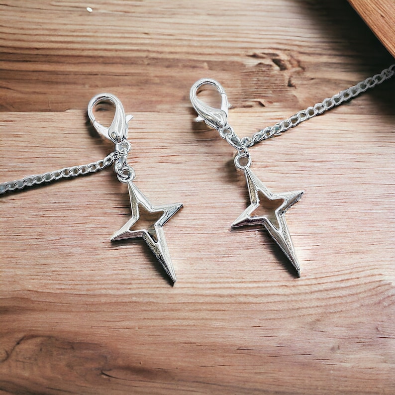 May include: Two silver star-shaped charms with a chain attached to each. The charms are hanging from a wooden surface.
