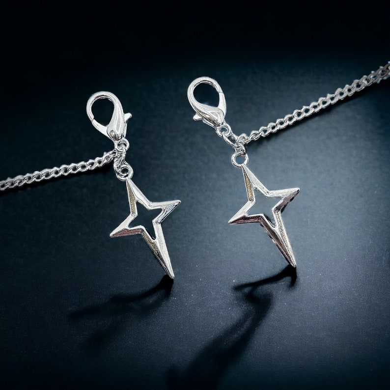 May include: Two silver star-shaped charms attached to a silver chain. The charms are dangling from the chain and are in focus.
