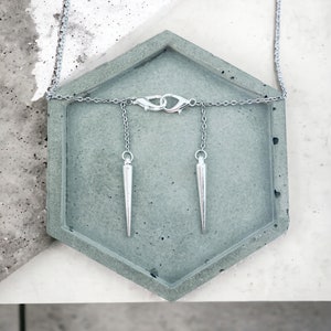 May include: A silver chain necklace with two silver pointed pendants hanging from it. The necklace is displayed on a gray concrete hexagonal tray.