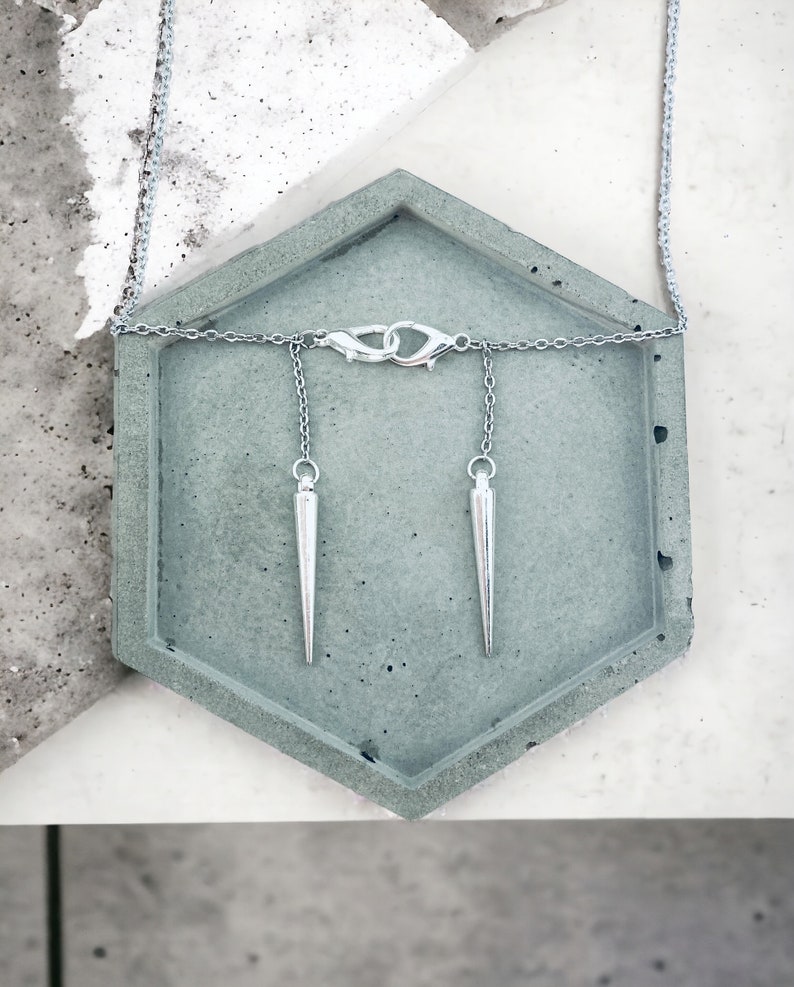 May include: A silver chain necklace with two silver pointed pendants hanging from it. The necklace is displayed on a gray concrete hexagonal tray.
