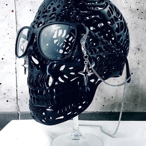 May include: A black skull sculpture with a detailed, intricate design. The skull is wearing round, black glasses with a silver chain attached to the side. The chain has a star-shaped charm.