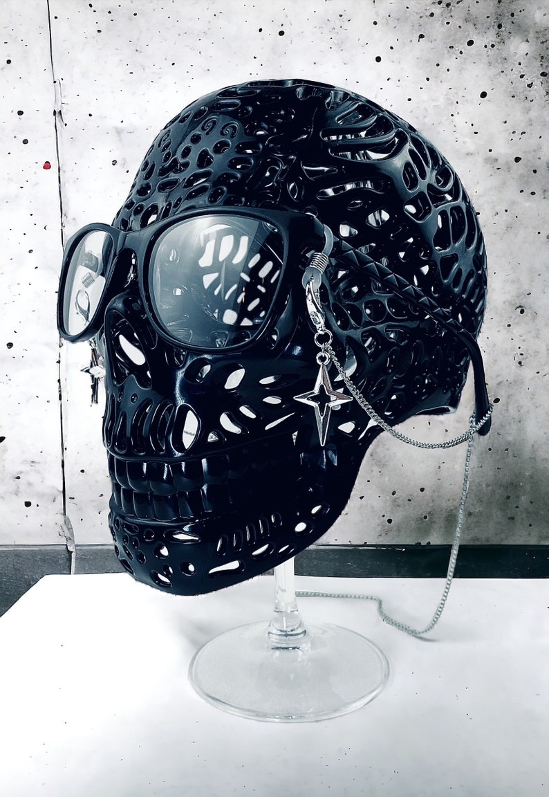 May include: A black skull sculpture with a detailed, intricate design. The skull is wearing round, black glasses with a silver chain attached to the side. The chain has a star-shaped charm.