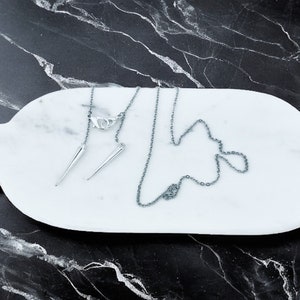 May include: A silver chain necklace with two silver spike pendants on a white marble surface.