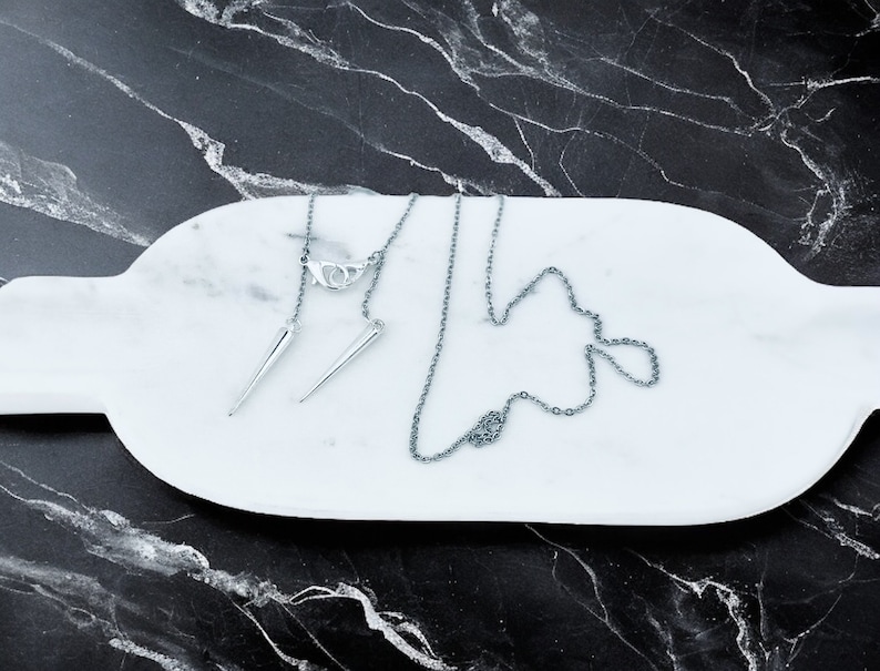 May include: A silver chain necklace with two silver spike pendants on a white marble surface.