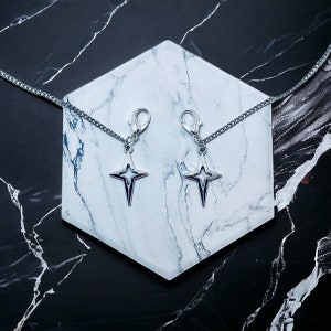 May include: Two silver star-shaped charms with a silver chain attached to each. The charms are hanging from a white marble surface.