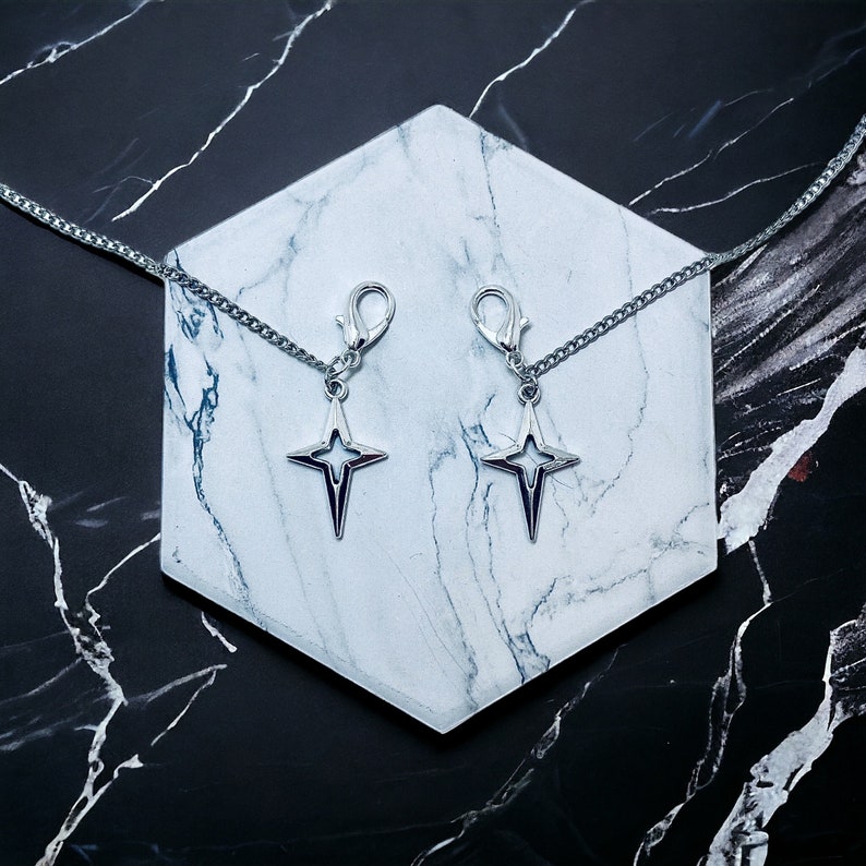 May include: Two silver star-shaped charms with a silver chain attached to each. The charms are hanging from a white marble surface.