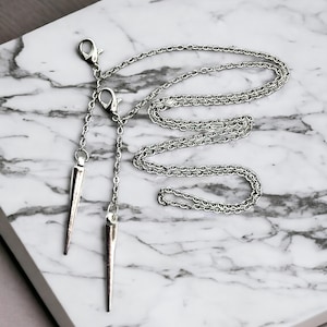 May include: A silver chain necklace with two sharp, pointed pendants. The necklace is on a white marble surface.