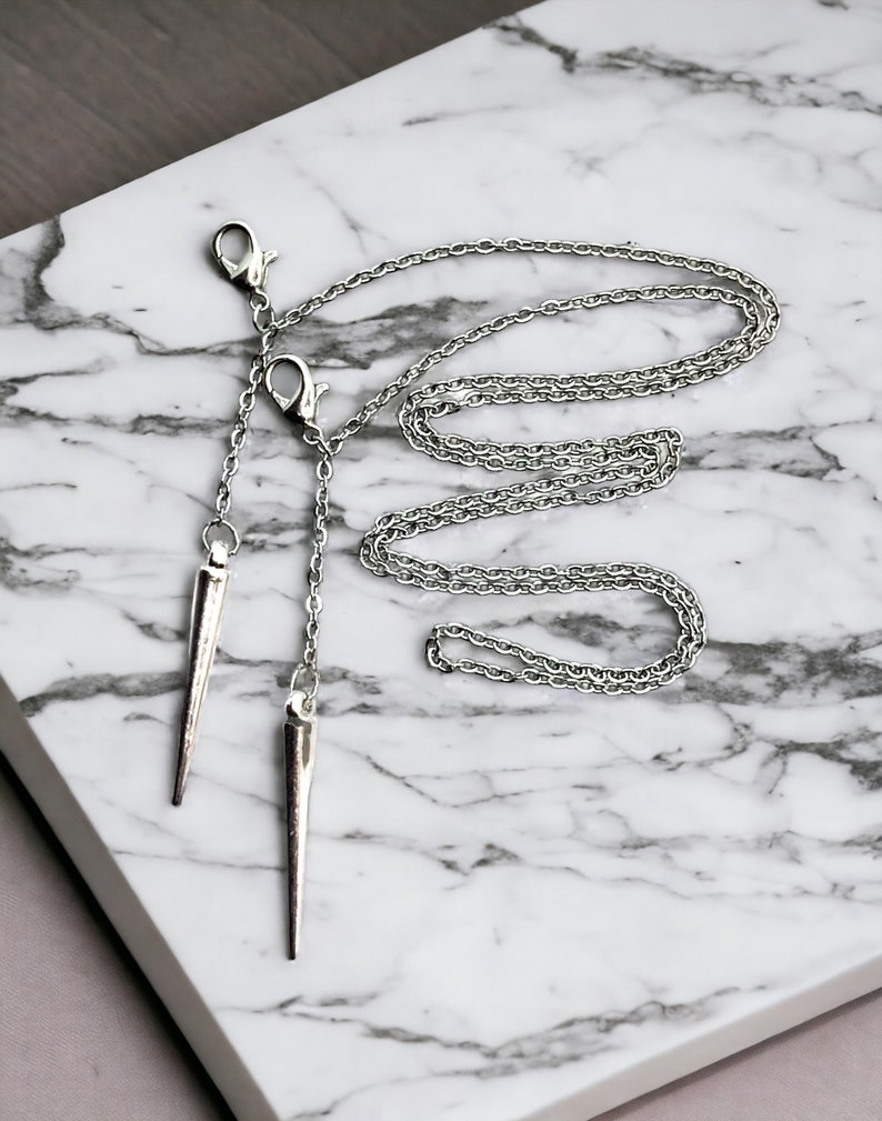 May include: A silver chain necklace with two sharp, pointed pendants. The necklace is on a white marble surface.