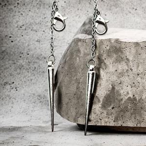 May include: Two silver chain earrings with sharp, pointed ends. The earrings are hanging from a concrete surface.