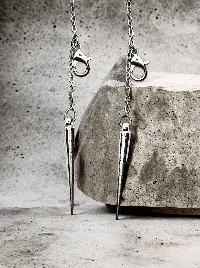 May include: Two silver chain earrings with sharp, pointed ends. The earrings are hanging from a concrete surface.