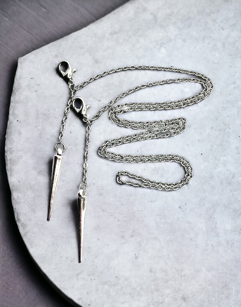 May include: A silver chain necklace with two sharp, pointed silver pendants hanging from it. The necklace is lying on a gray surface.