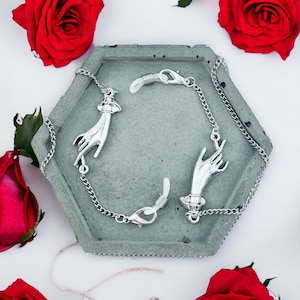 May include: A pair of silver eyeglass chain holders with hand-shaped charms. The chains are on a gray hexagonal surface with red roses in the background.