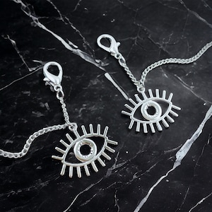May include: Two silver eye-shaped charms on a silver chain with a lobster clasp. The eyes have a detailed design with lashes and a circle in the center.