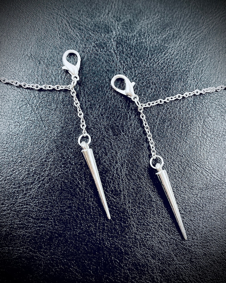 May include: Two silver chains with sharp, pointed metal tips hanging from silver clasps. The chains are against a black background.