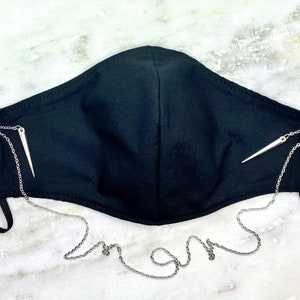May include: Black face mask with a silver chain and two silver spikes attached to the straps. The chain is long enough to hang around the neck.