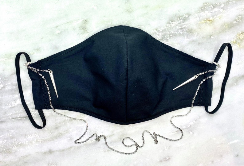 May include: Black face mask with a silver chain and two silver spikes attached to the straps. The chain is long enough to hang around the neck.