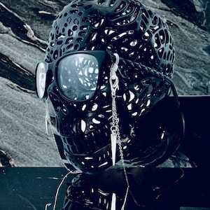 May include: A black skull-shaped object with a detailed, intricate design. The skull is wearing sunglasses and has two silver chains with pointed pendants hanging from the ears.