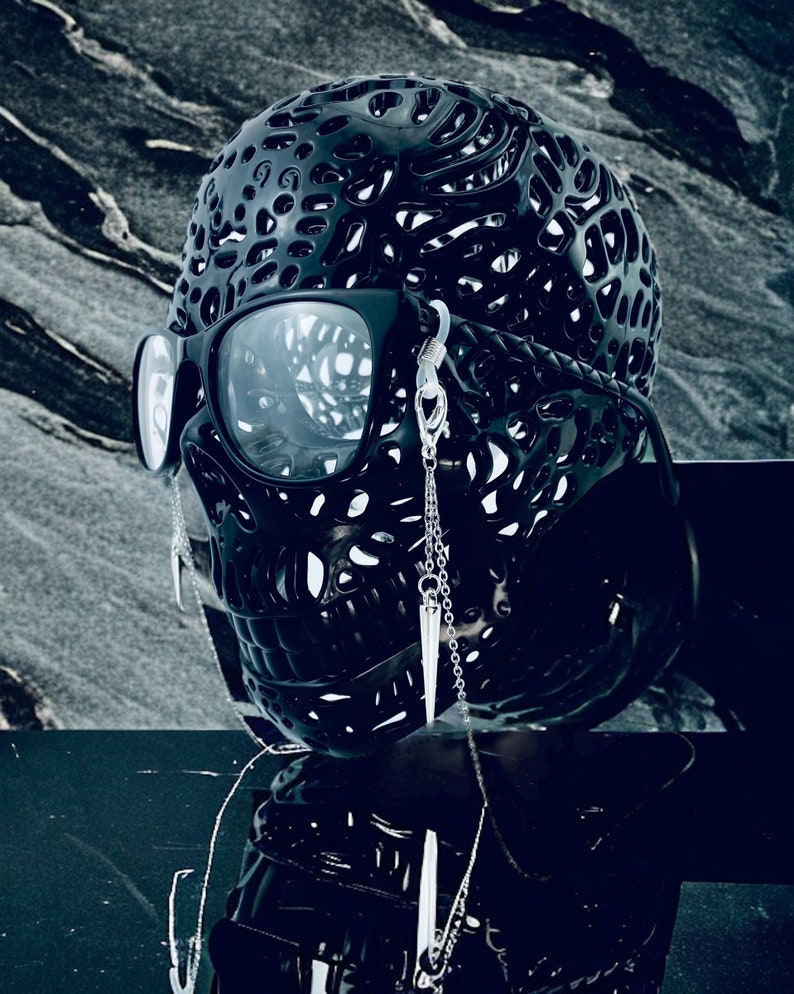 May include: A black skull-shaped object with a detailed, intricate design. The skull is wearing sunglasses and has two silver chains with pointed pendants hanging from the ears.