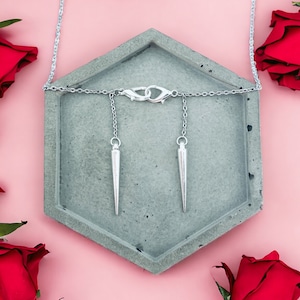 May include: A silver necklace with two sharp, pointed pendants hanging from it. The necklace is displayed on a gray hexagonal tray, surrounded by red roses.