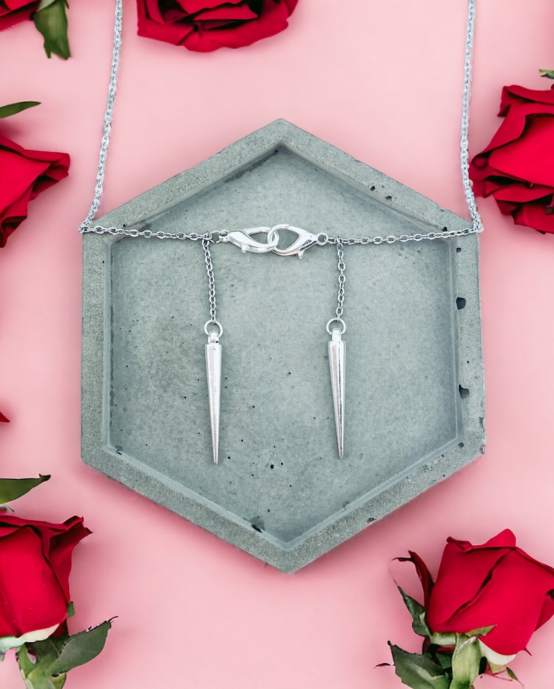 May include: A silver necklace with two sharp, pointed pendants hanging from it. The necklace is displayed on a gray hexagonal tray, surrounded by red roses.