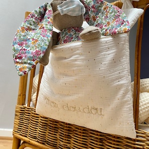 Peut inclure: Un sac de rangement en tissu blanc avec des paillettes dorées et le texte "mon doudou" brodé dessus. Un lapin en peluche avec des oreilles en tissu fleuri est à l'intérieur du sac. Le sac est posé sur un panier en osier.