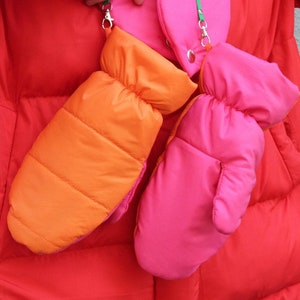 Puede incluir: Un par de guantes de color naranja brillante y rosa con una correa verde unida a cada uno. Los guantes están colgando de una chaqueta roja acolchada.
