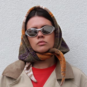 May include: A person wearing a beige jacket with a brown corduroy collar, a red shirt, and a multi-colored scarf tied around their head. They are wearing oval-shaped sunglasses.