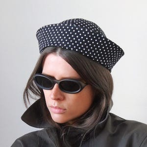 May include: A black beret with a white polka dot pattern, paired with dark oval-shaped sunglasses and a dark olive green trench coat featuring a high collar and visible buttons on the sleeves, creating a structured and layered appearance.
