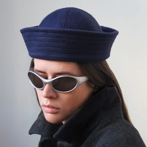May include: A navy blue sailor hat with a structured brim. The hat is worn with silver-framed sunglasses and a dark gray coat. The image is set against a plain, light gray background.