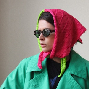 May include: A woman wearing a green trench coat with a pink and green knitted scarf. The scarf is tied around her head and neck.