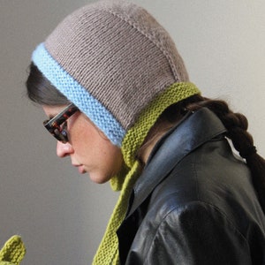 Könnte beinhalten: Eine Frau trägt eine braune Strickmütze mit blauem und grünem Besatz, eine schwarze Lederjacke und einen grünen Strickschal.