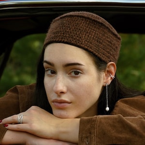May include: A woman wearing a brown knitted headband, a suede jacket, and a silver ring. A silver earring with a dangling pearl is visible. The woman is leaning on a red car.